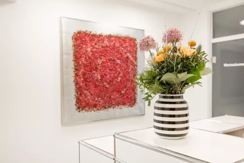 Ein Blumenstrauß in einer gestreiften Vase steht auf dem Empfangstresen. Im Hintergrund hängt eine rote Kunstinstallation an der Wand.